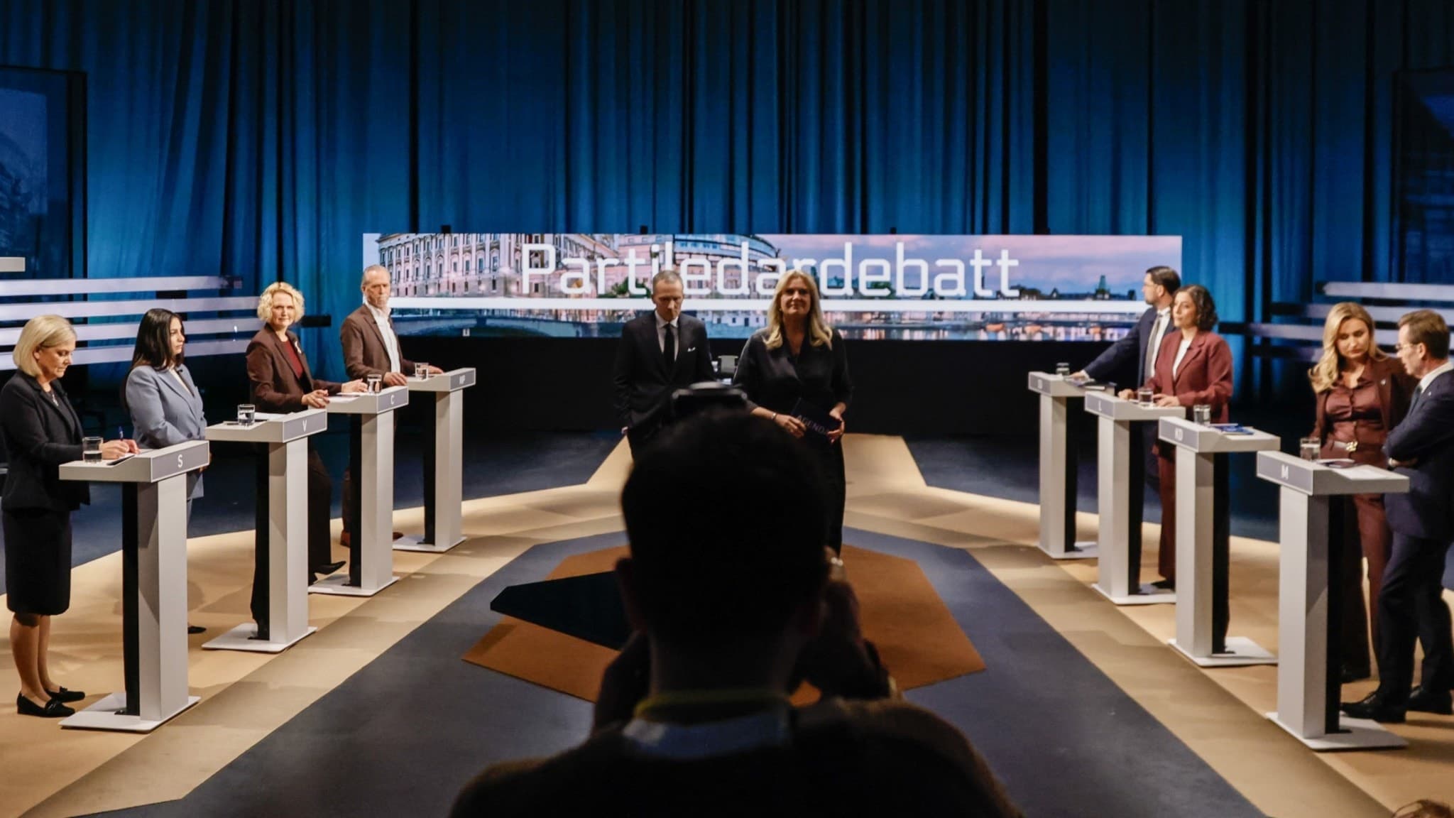 Riksdagens åtta partiledare står vid sina podier i en TV-studio under SVT:s Agenda. Programledarna Anders Holmberg och Camilla Kvartoft syns i mitten. Studion är upplyst med blått ljus.